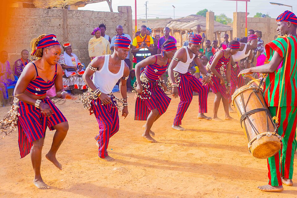 A group of dancers in matching red and blue striped traditional dress, mid-performance beside a drummer in Ghana